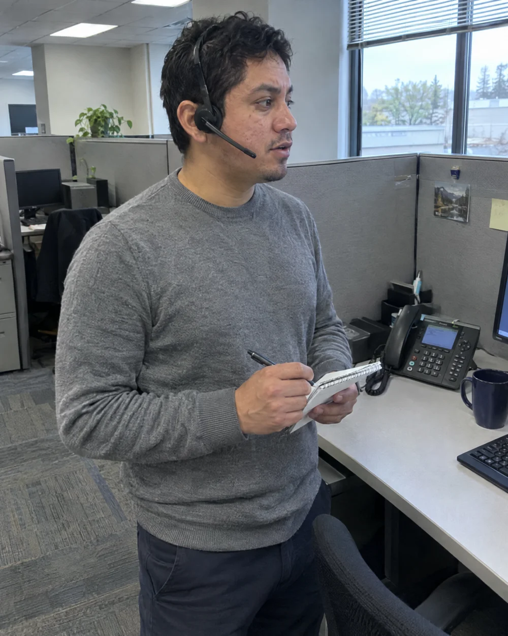 Recruiter on a headset call with a client, holding his notes beside an office window